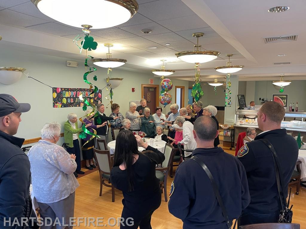 A festive gathering in a dining hall, celebrating Sr. Jo's birthday with seniors, decorations, and firefighters present.
