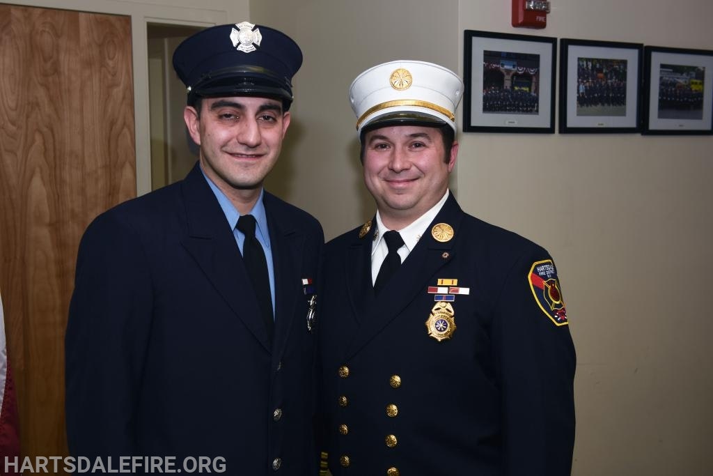 Two firefighters in uniform smiling, standing indoors with framed photos on the wall behind them.