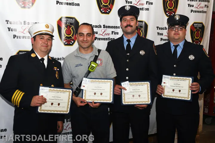 Four firefighters with awards at a recognition event, standing in front of a Hartsdale Fire District banner.