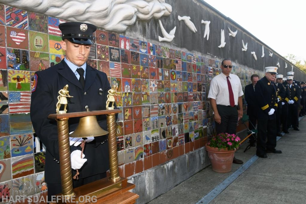 A firefighter holds a ceremonial bell in front of a mural with onlookers standing in a line, likely during a memorial event.