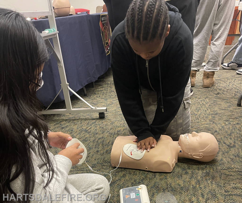 A child is practicing CPR on a manikin while another child prepares to use an AED in a training session.