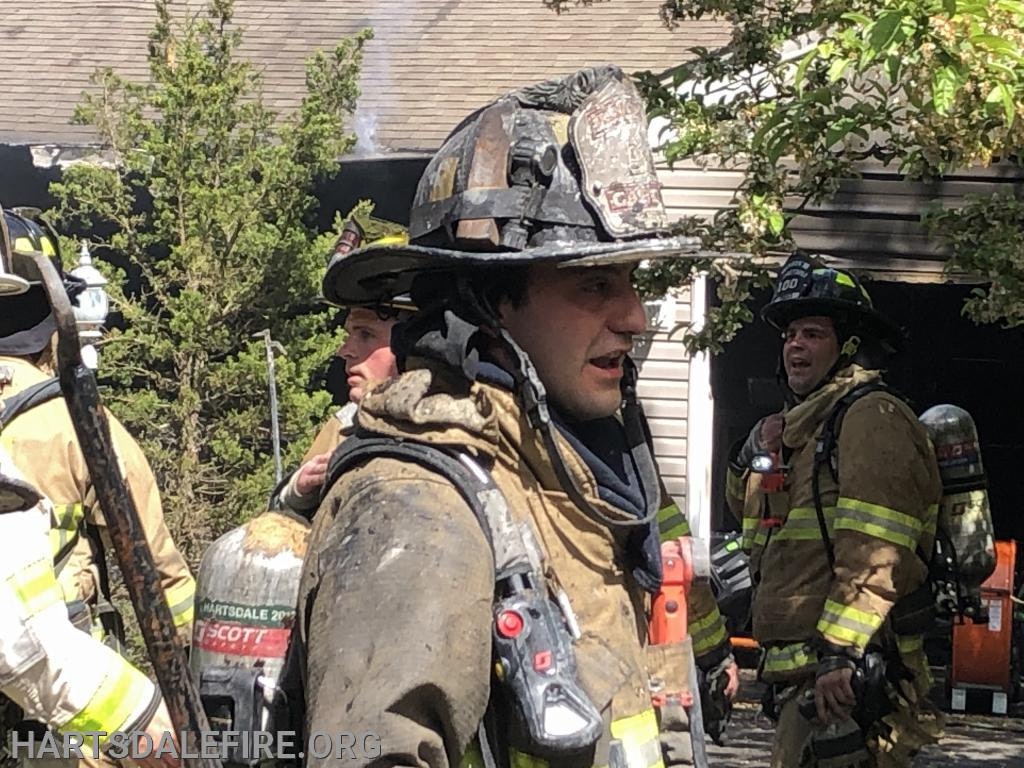 Firefighters in gear outside a building with smoke, trees, and equipment visible.