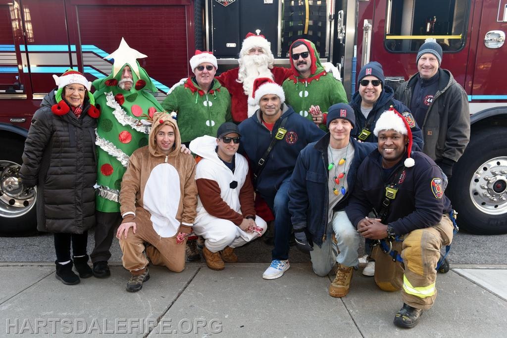 A festive group of people, including firefighters in holiday attire, posing in front of a fire truck for a cheerful photo.