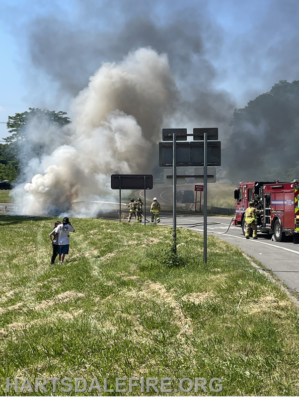 A smoky scene shows firefighters responding to a fire near a roadway, while bystanders observe from a distance.