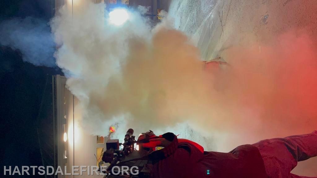 A person in firefighting gear using a smoke machine outdoors at night.