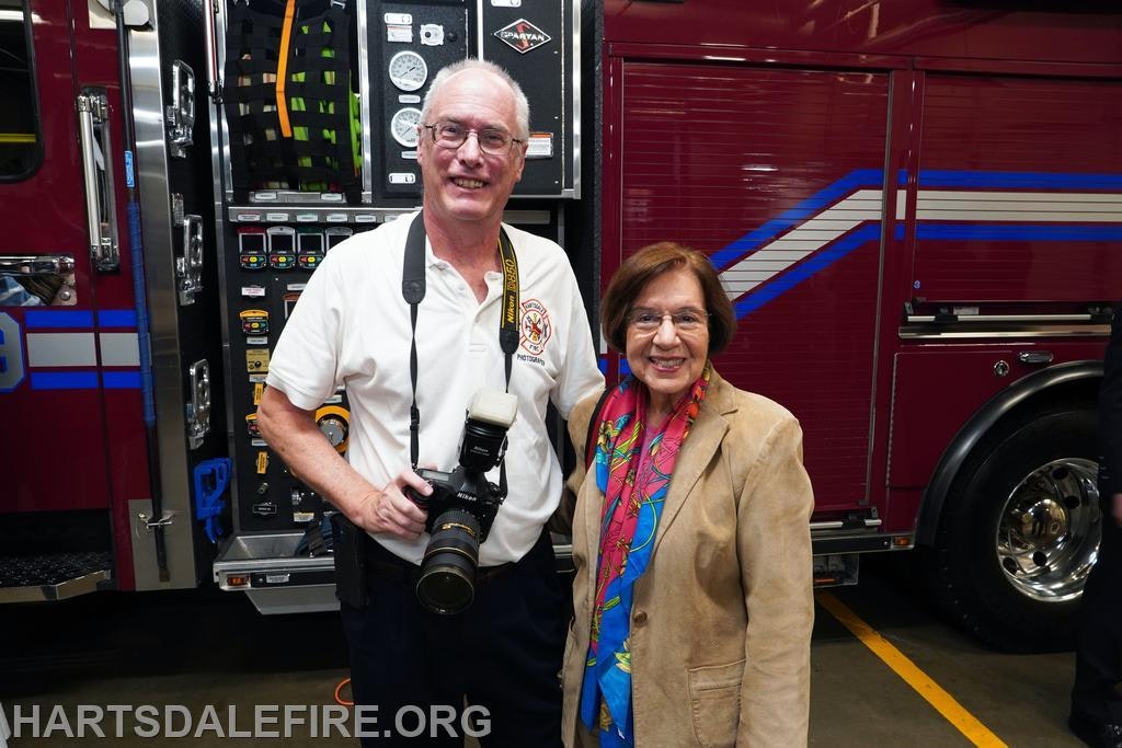 Two people smiling, one holding a camera, in front of a fire truck with open equipment panel.