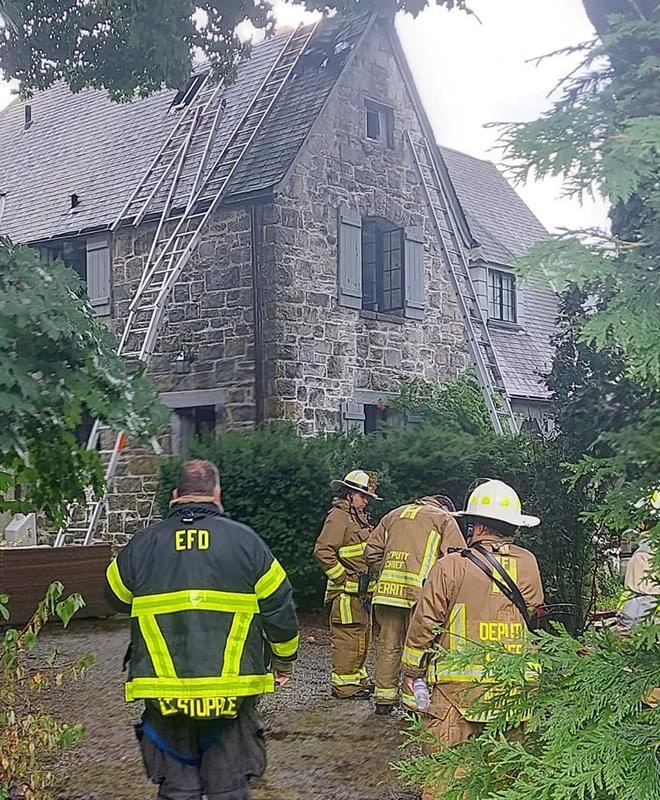 Firefighters in gear outside a stone house with ladders on the roof.
