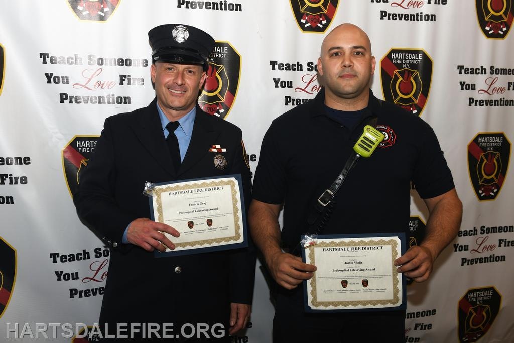 Two firefighters holding awards in front of a Hartsdale Fire District backdrop.