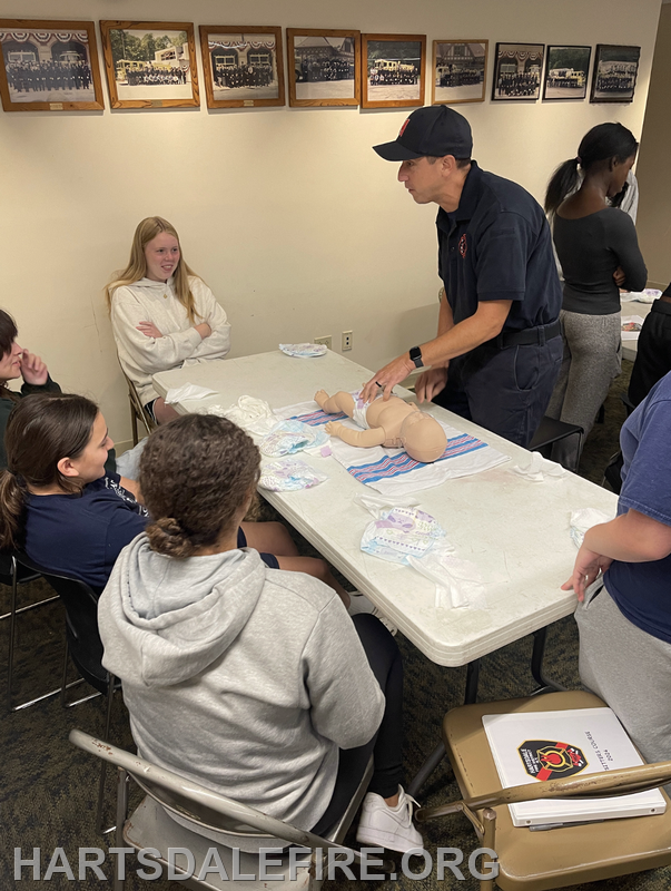 A fire instructor teaches a group about infant care using a doll in a classroom setting.