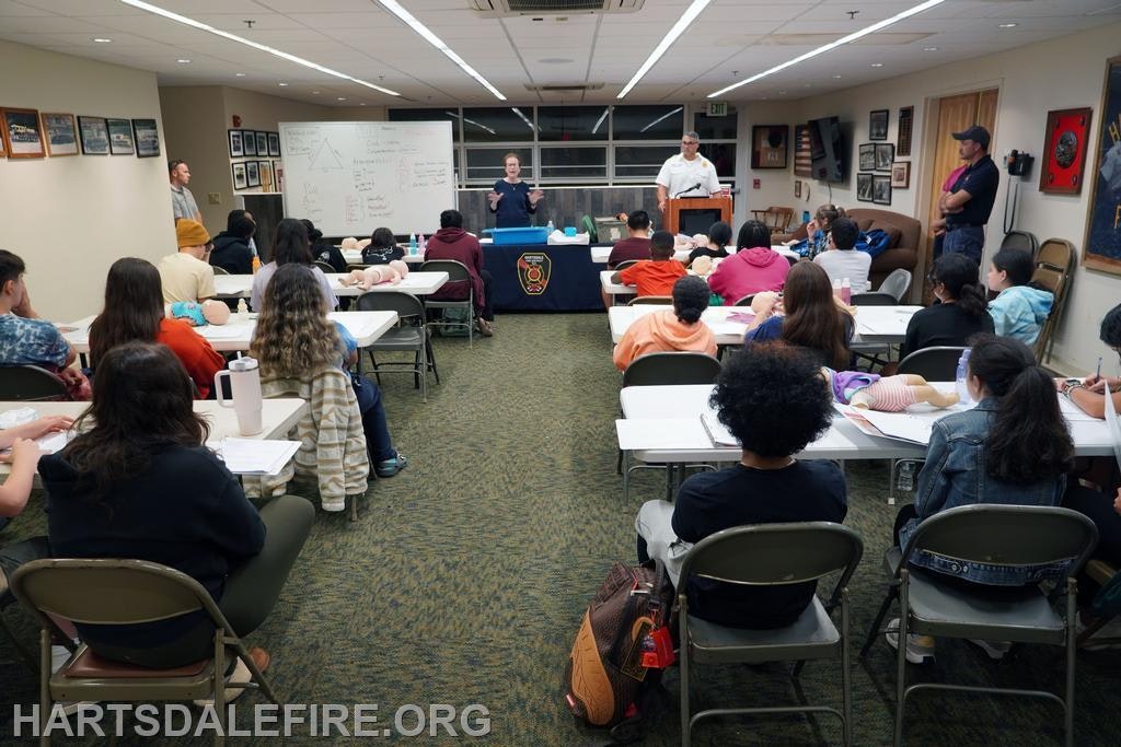 Classroom with students attending a lecture or training session, with mannequins on tables, and two instructors at the front.