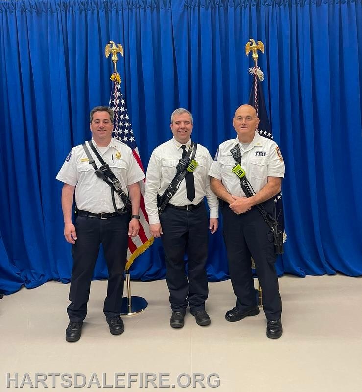 Three firefighters in uniforms stand in front of flags and blue curtains.