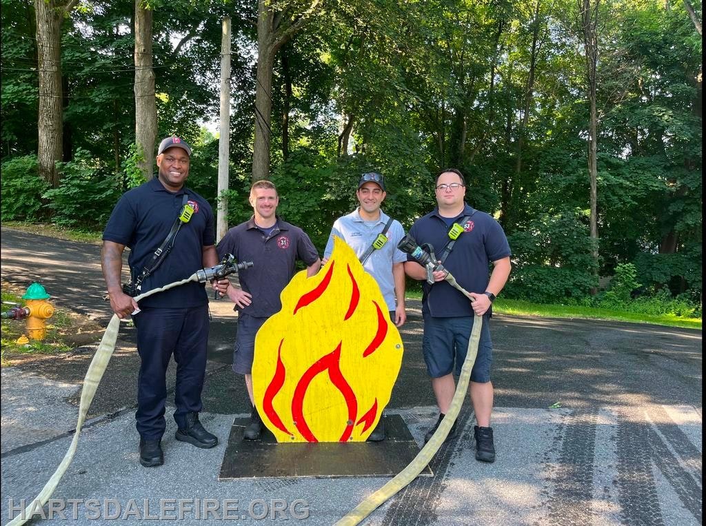 Four people pose with a fire hose and a cutout of a flame on a road. Trees and a fire hydrant are in the background.