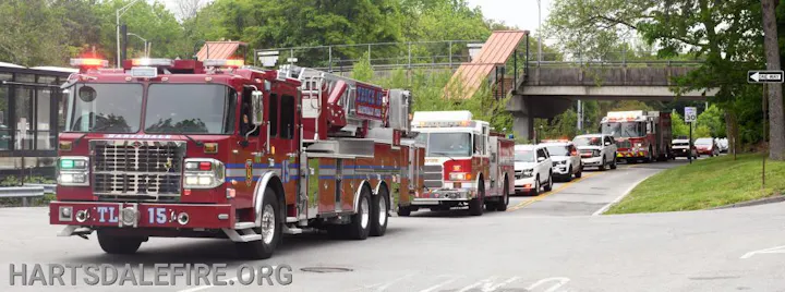 A lineup of fire trucks and emergency vehicles on a road near a bridge, trees in the background.