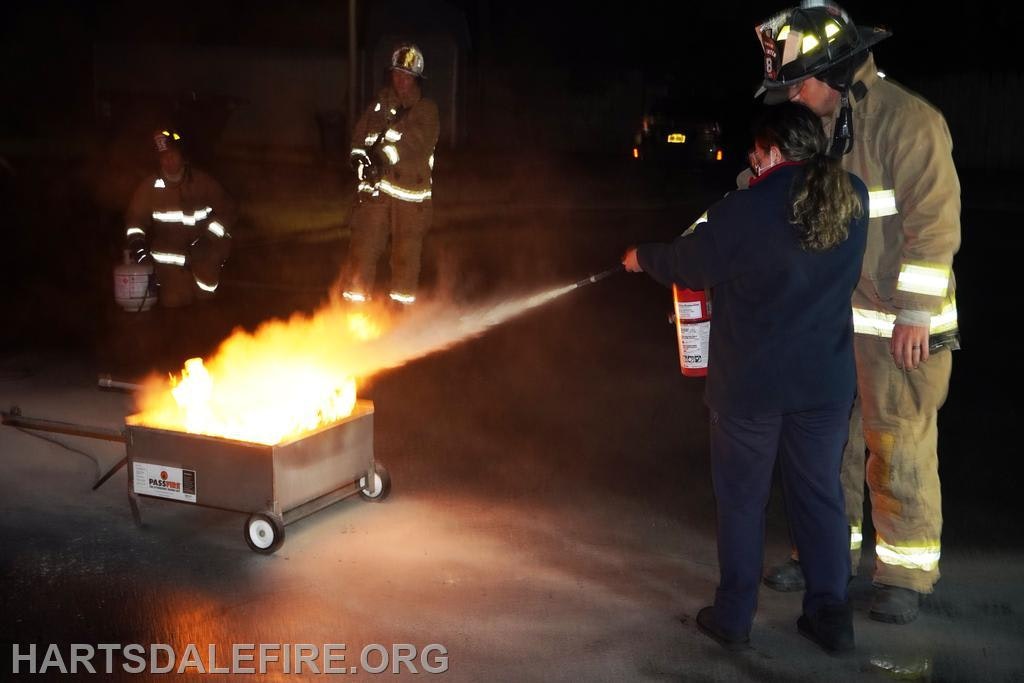 A firefighter supervises as a person uses a fire extinguisher on a controlled fire during training.
