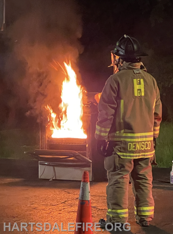 A firefighter stands by a burning object at night, observing the flames and smoke, ready to act.