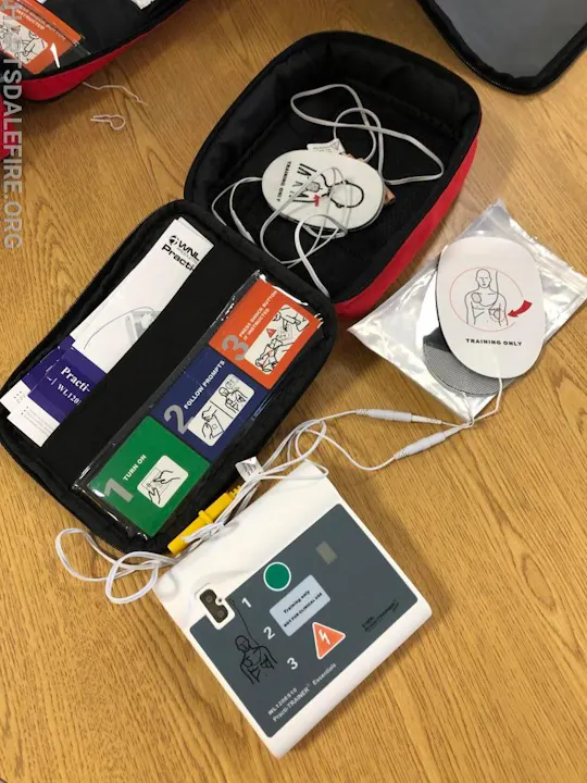 A defibrillator training kit with pads, manuals, and labeled instructions on a wooden surface.