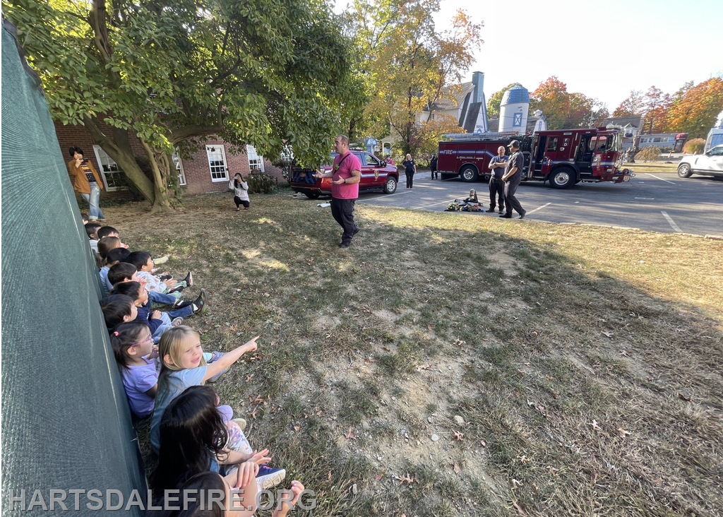 Children watch a demonstration by firefighters near a fire truck, with some adults observing in the background.