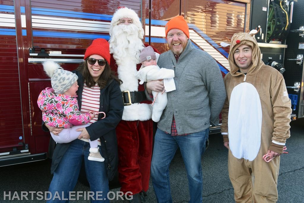A festive group poses with Santa and a reindeer, holding babies, in front of a firetruck. Holiday cheer abounds!