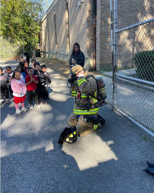 A firefighter kneels to engage with a group of children watching, while an adult stands nearby.