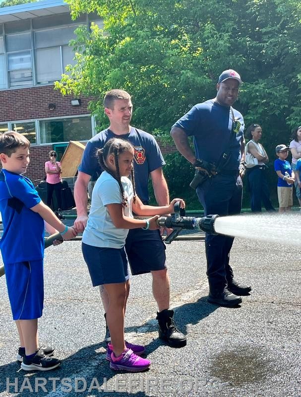 Children and firefighters use a hose to spray water during an outdoor event or demonstration.