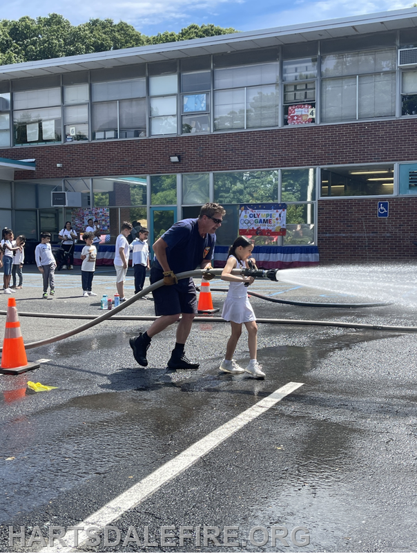A firefighter and a young girl are spraying water from a fire hose at an outdoor school event with children watching nearby.
