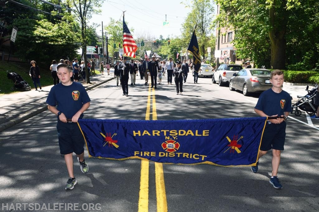 A parade with people holding a "Hartsdale Fire District" banner, followed by uniformed individuals carrying flags.