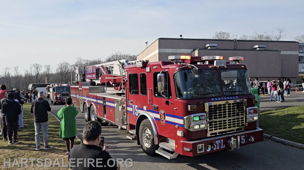 A red fire truck with lights is parked, while people gather and watch in a school setting under clear skies.
