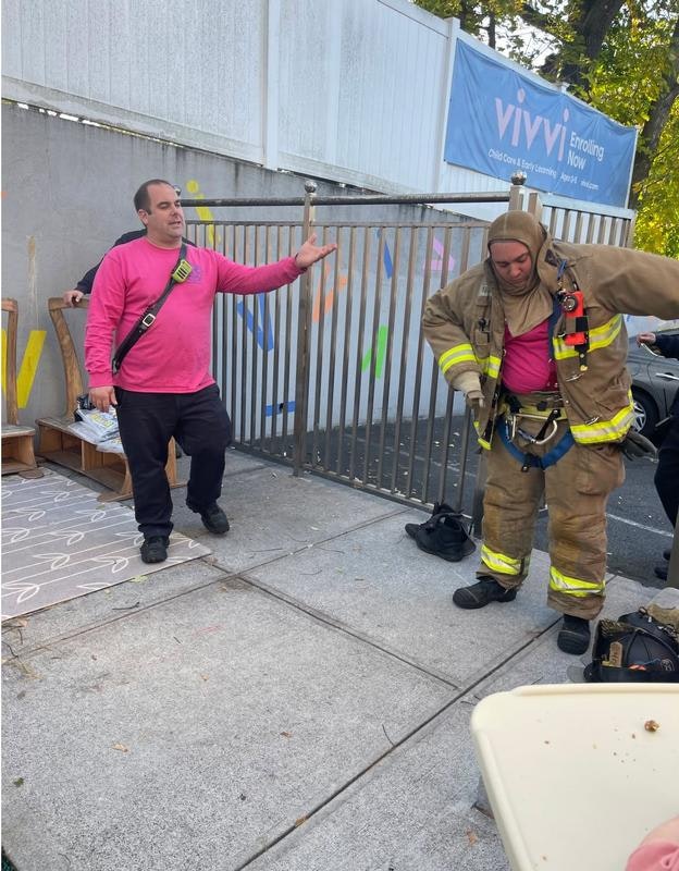 The image shows two men: one in a pink shirt and the other in firefighter gear, preparing for an activity near a fence.