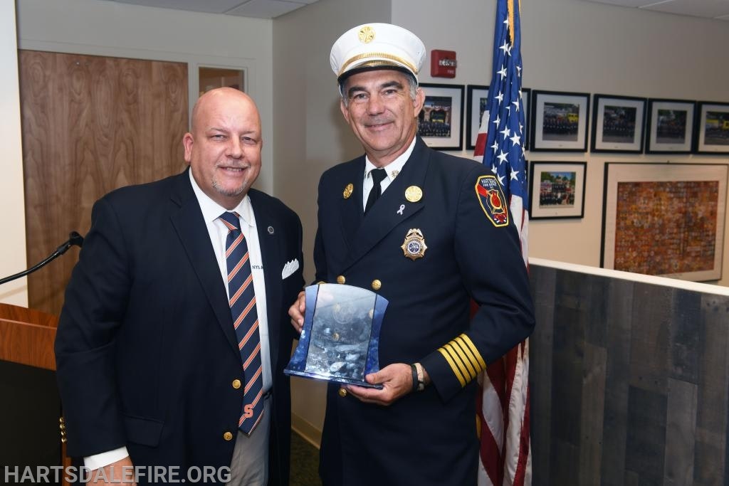 Two men pose for a photo, one in a suit, the other in a fire chief uniform holding an award, in front of an American flag.