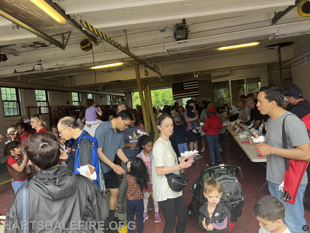 A crowded fire station event with people, including families and children, enjoying food and activities. An American flag is visible.