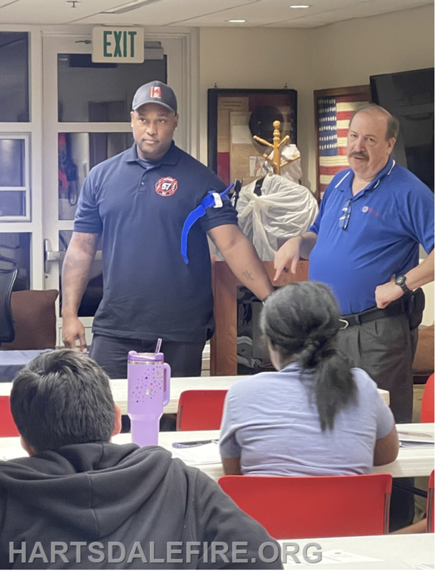 Two individuals, one in a blue shirt and another in a black shirt, speak to a group in a classroom setting.