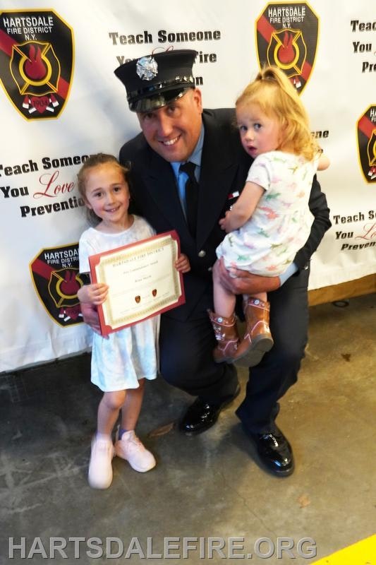 A firefighter kneels, smiling with two children. One child holds a certificate, and a fire safety banner is in the background.