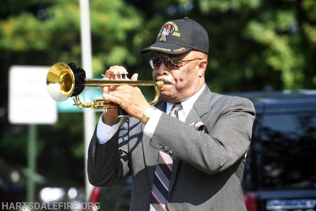 A man in a suit plays a trumpet outdoors, wearing a hat and sunglasses.