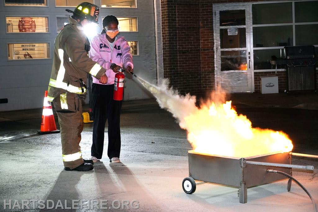 A firefighter helps a person use a fire extinguisher to put out a controlled fire in a metal pan.