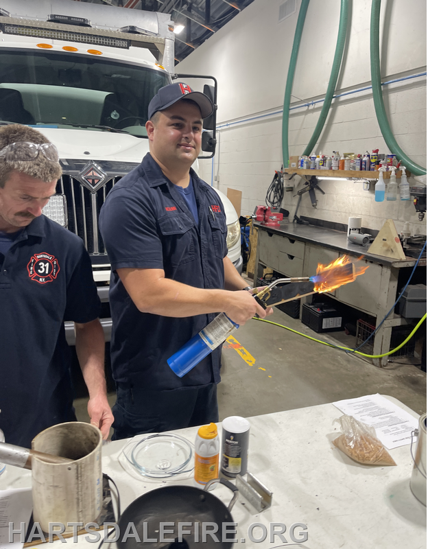 A firefighter is using a propane torch with a flame, while another person looks on in a workshop with various tools and chemicals.