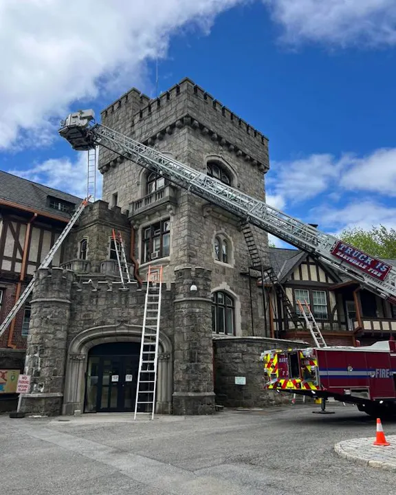 A fire truck with a ladder is positioned near a stone tower building, with a blue sky in the background.