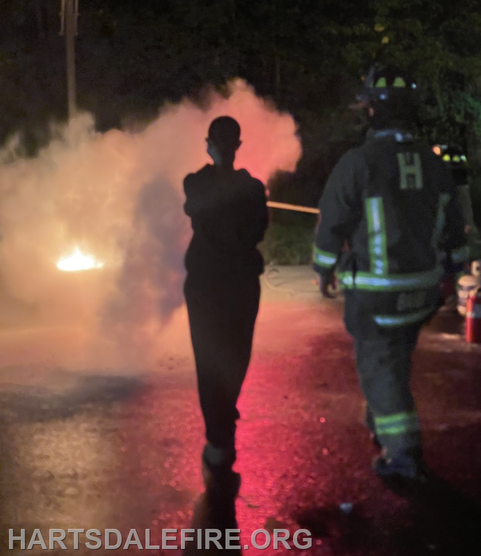 A firefighter approaches a smoky scene at night, where a small fire appears to be present. Safety precautions are in place.