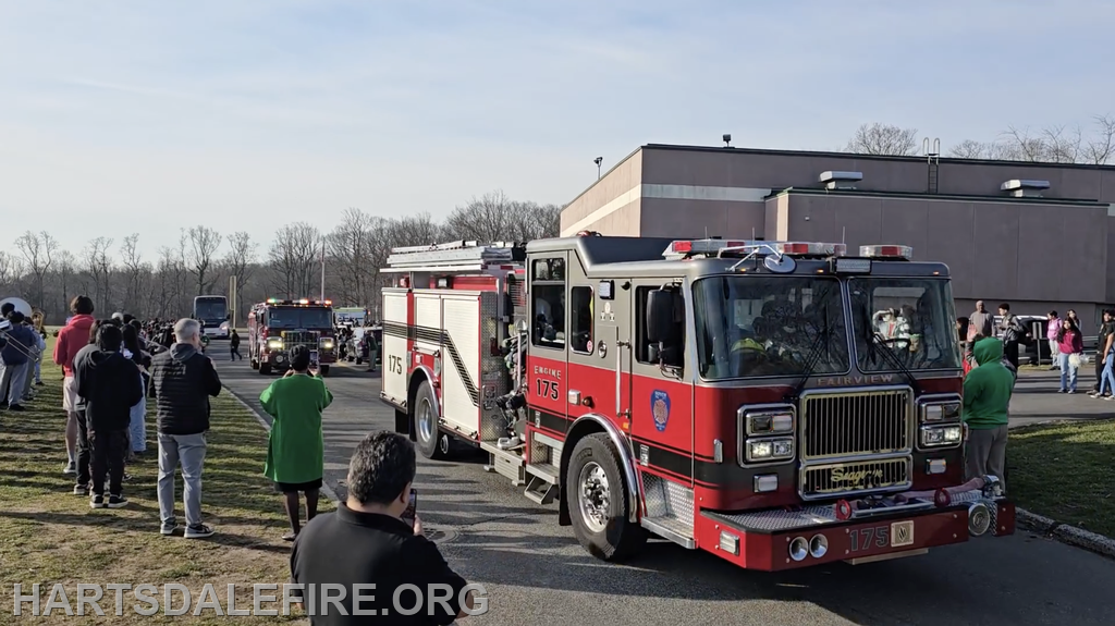 A fire truck is parading past a crowd of onlookers outside a building on a clear day.