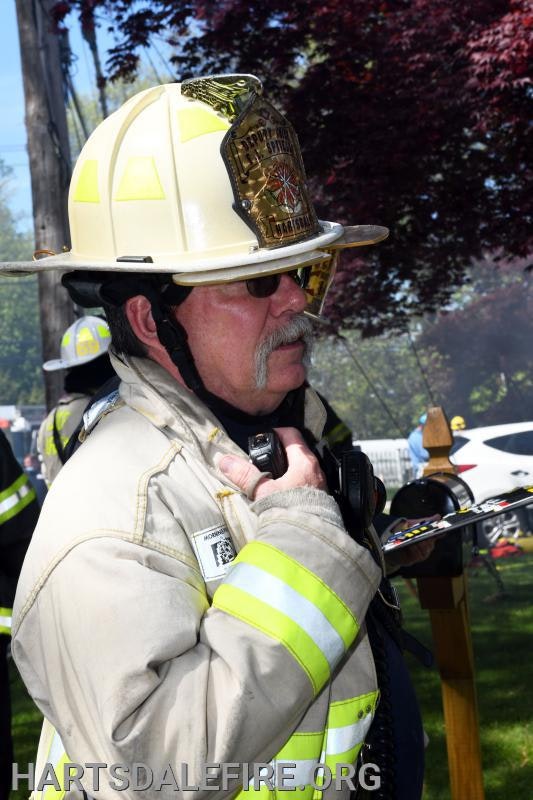 A firefighter in full gear holding a radio, standing outside, with trees and a car nearby.