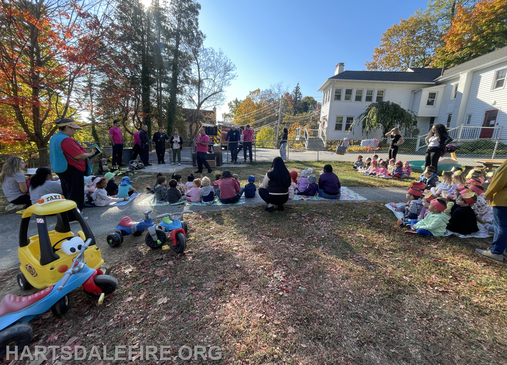 A group of children is sitting outside, watching a performance by adults, with colorful trees and toys nearby.