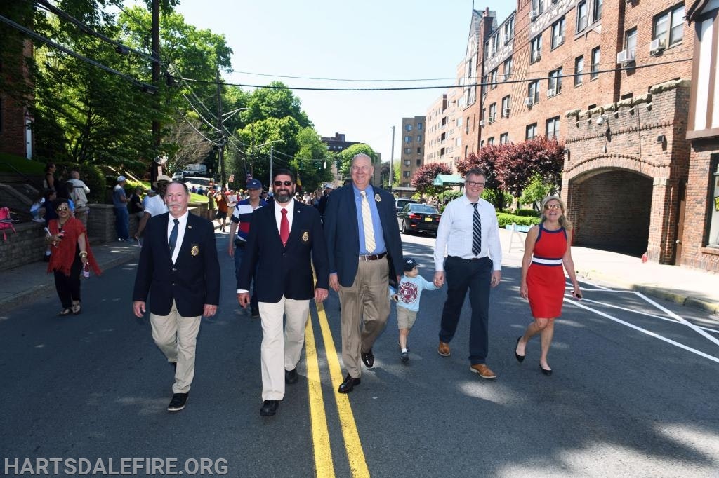 A group of people in formal attire walking on a street during a parade or event, with buildings and onlookers in the background.