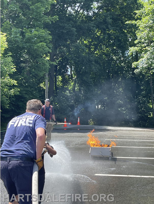 A firefighter uses a hose to extinguish a small fire while another individual observes, surrounded by cones in a wooded area.