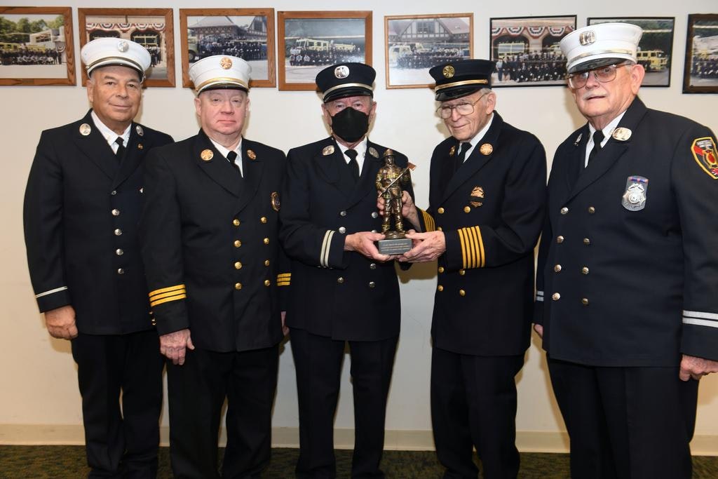 Five men in uniform, one holding a firefighter statue award, stand in front of framed photo displays on the wall.