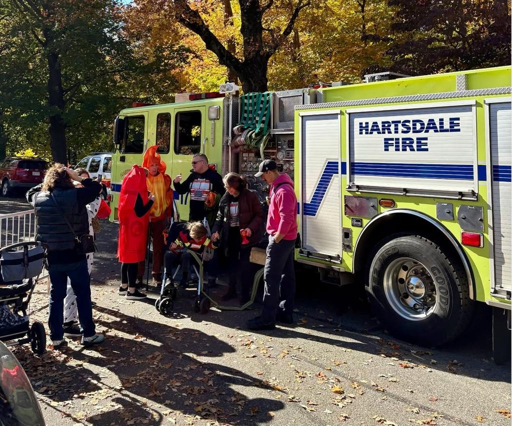 A group of people in costumes gathers around a Hartsdale fire truck on an autumn day.
