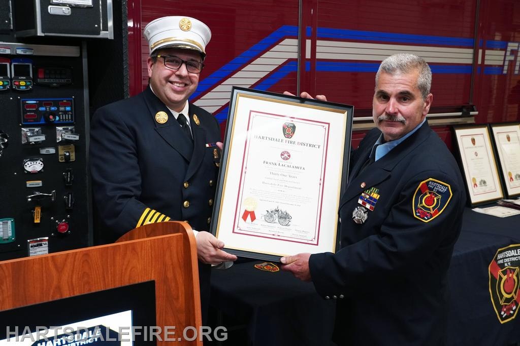 Two firefighters holding a framed certificate, standing in front of a fire truck.