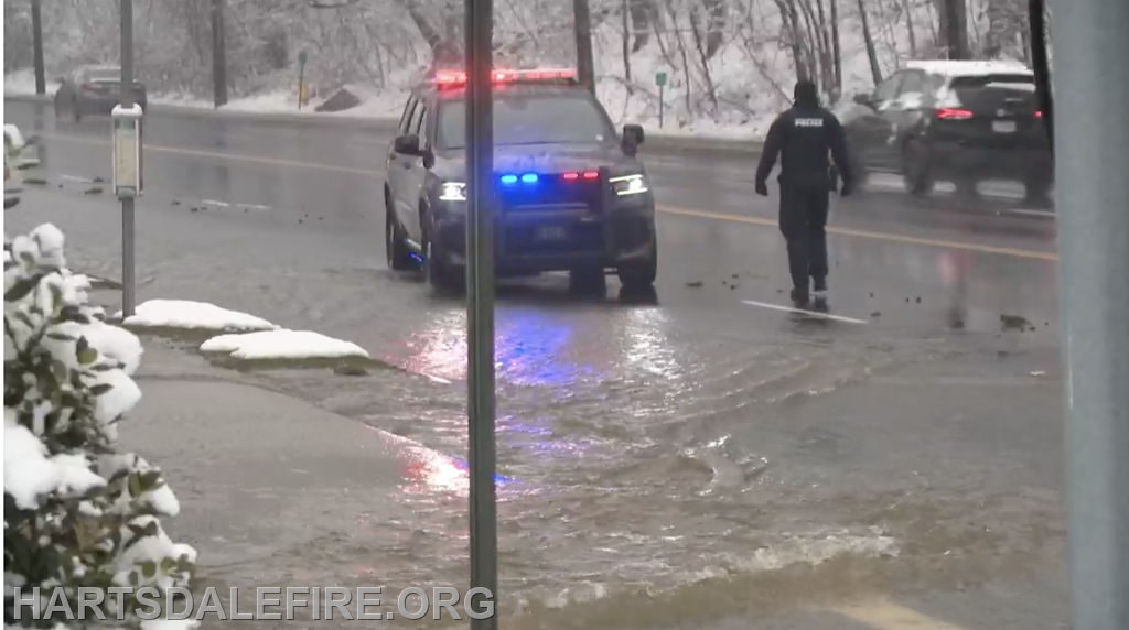 A police vehicle with lights on is parked on a flooded road, while an officer walks nearby, and snow covers the landscape.