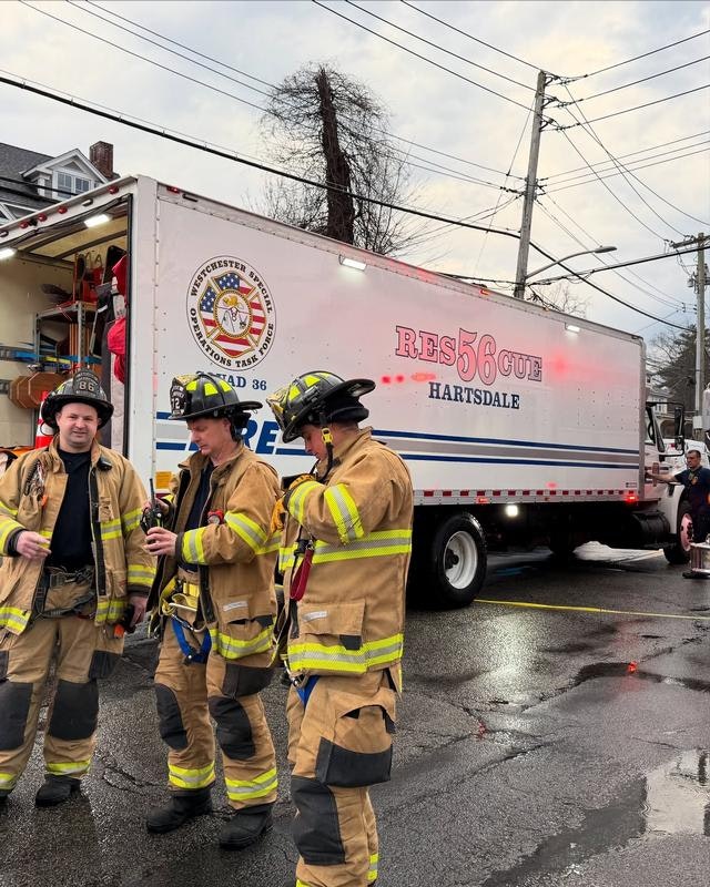 Firefighters in uniform are gathered near a rescue truck labeled "Hartsdale." The scene is wet and urban, likely responding to an incident.