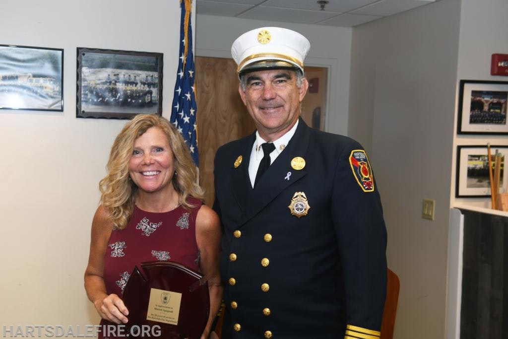 A woman with an award and a smiling firefighter in uniform stand together in a room with flags and framed pictures.