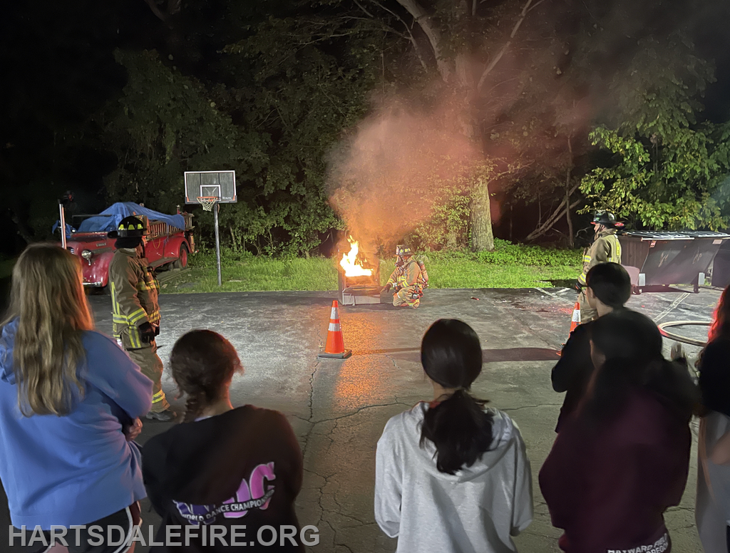 Firefighters conduct a demonstration for a group, showcasing fire safety techniques at night. An open flame burns in the background.