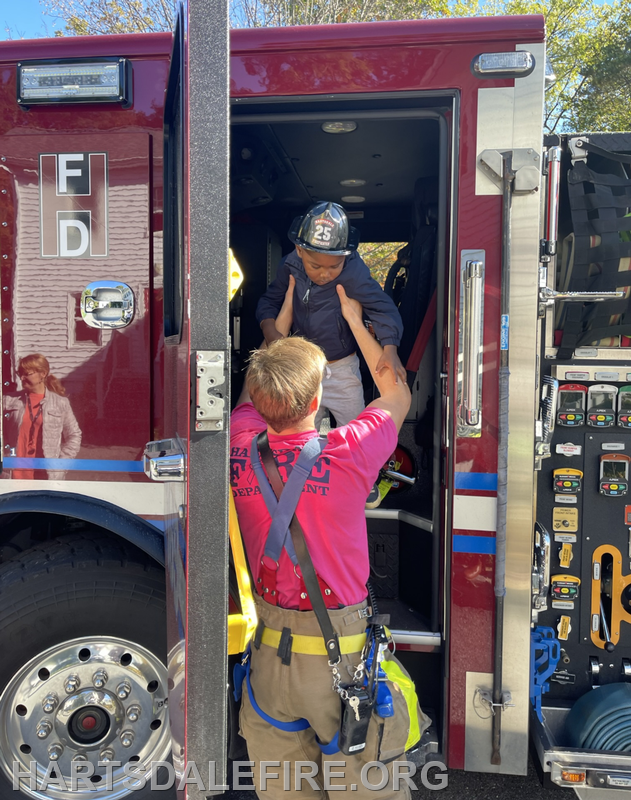 A firefighter helps a child in a helmet get into a fire truck, showcasing a community event or safety demonstration.